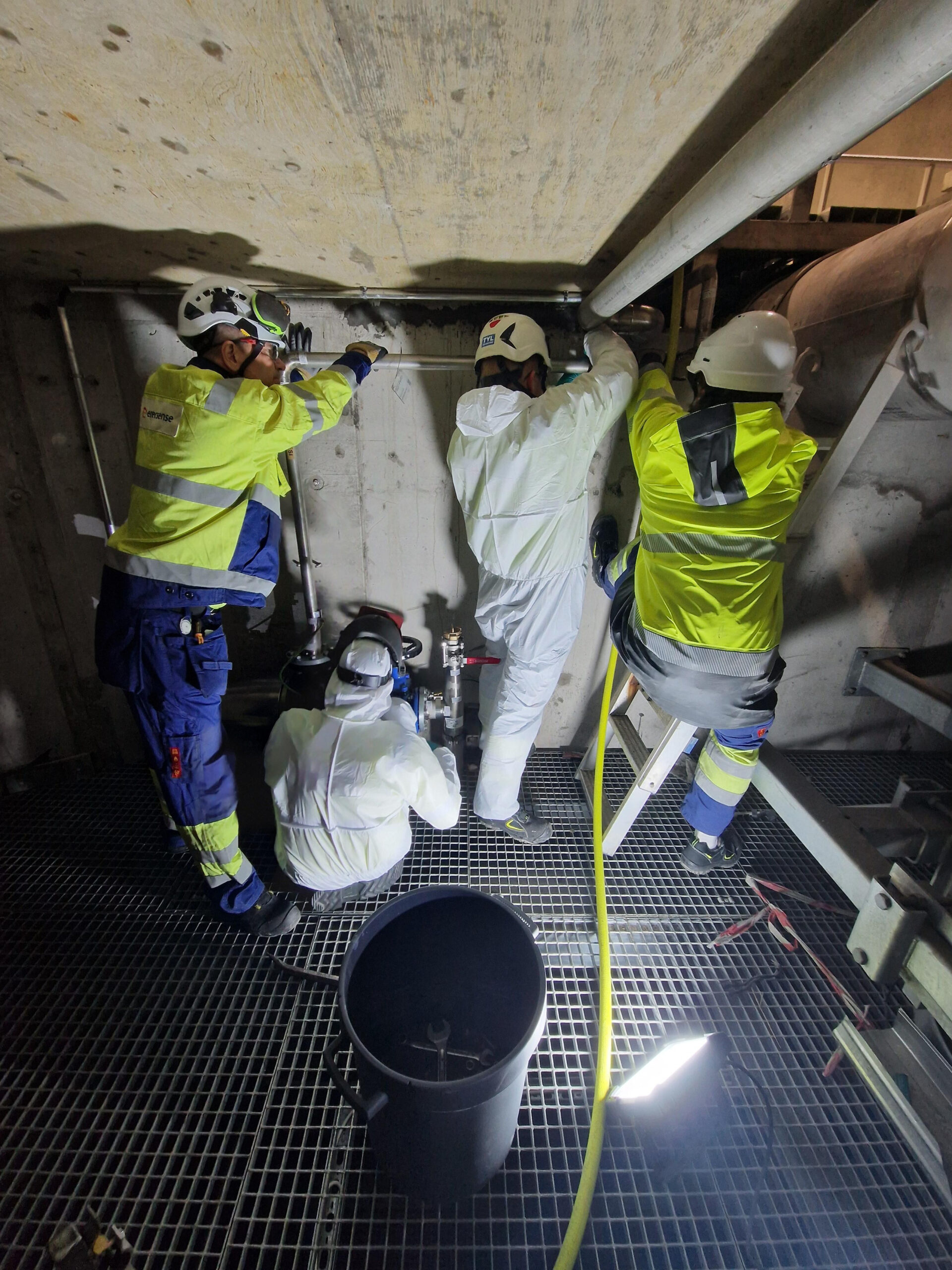 Wearing disposable protective coveralls, or Tyvek suits, the fitters reinstall a pipe at the Sulkavuori central treatment plant, assisted by two electricians handling the rigid pipe.